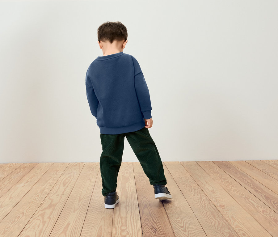 Ein Junge steht mit dem Rücken zum Betrachter und trägt ein blaues Sweatshirt und grüne Hosen.