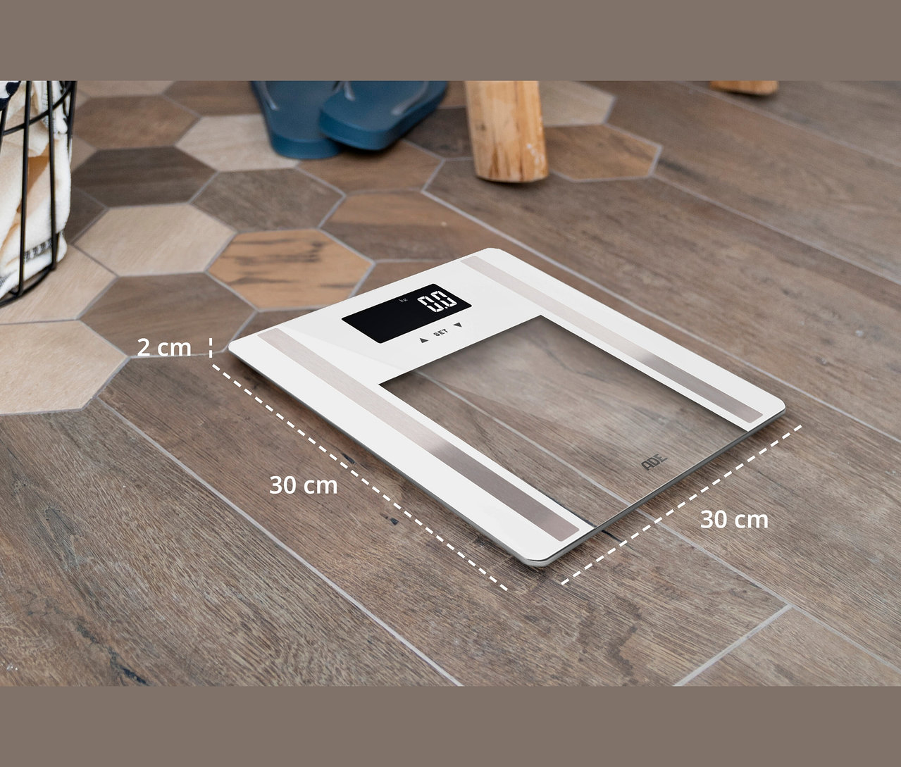 Eine weiße digitale ADE Personenwaage liegt auf einem braunen Boden. Die Waage ist 30 x 30 cm groß und 2 cm dick.