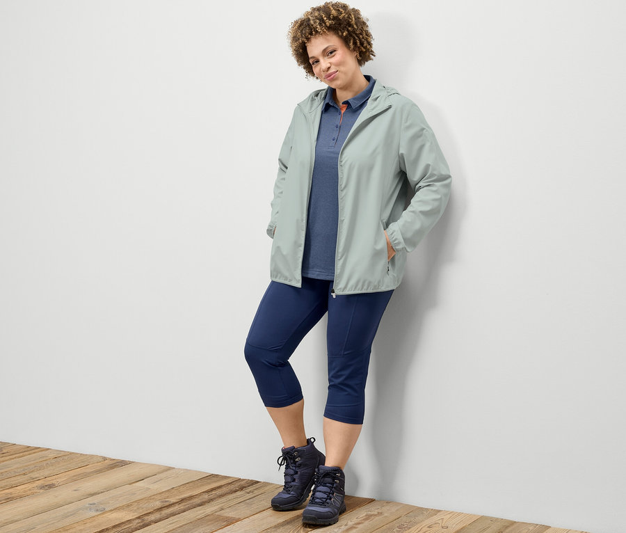 Eine Frau lehnt an einer Wand und trägt eine 3/4-Trekking-Tight, ein dunkelblaues Funktions-Poloshirt und eine graue, packbare Funktionsjacke.