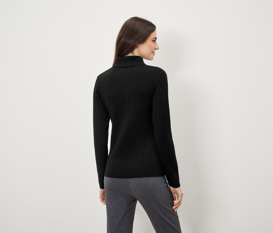 Frau mit schwarzem Rollkragenpullover und grauer Hose steht mit dem Rücken zur Kamera.