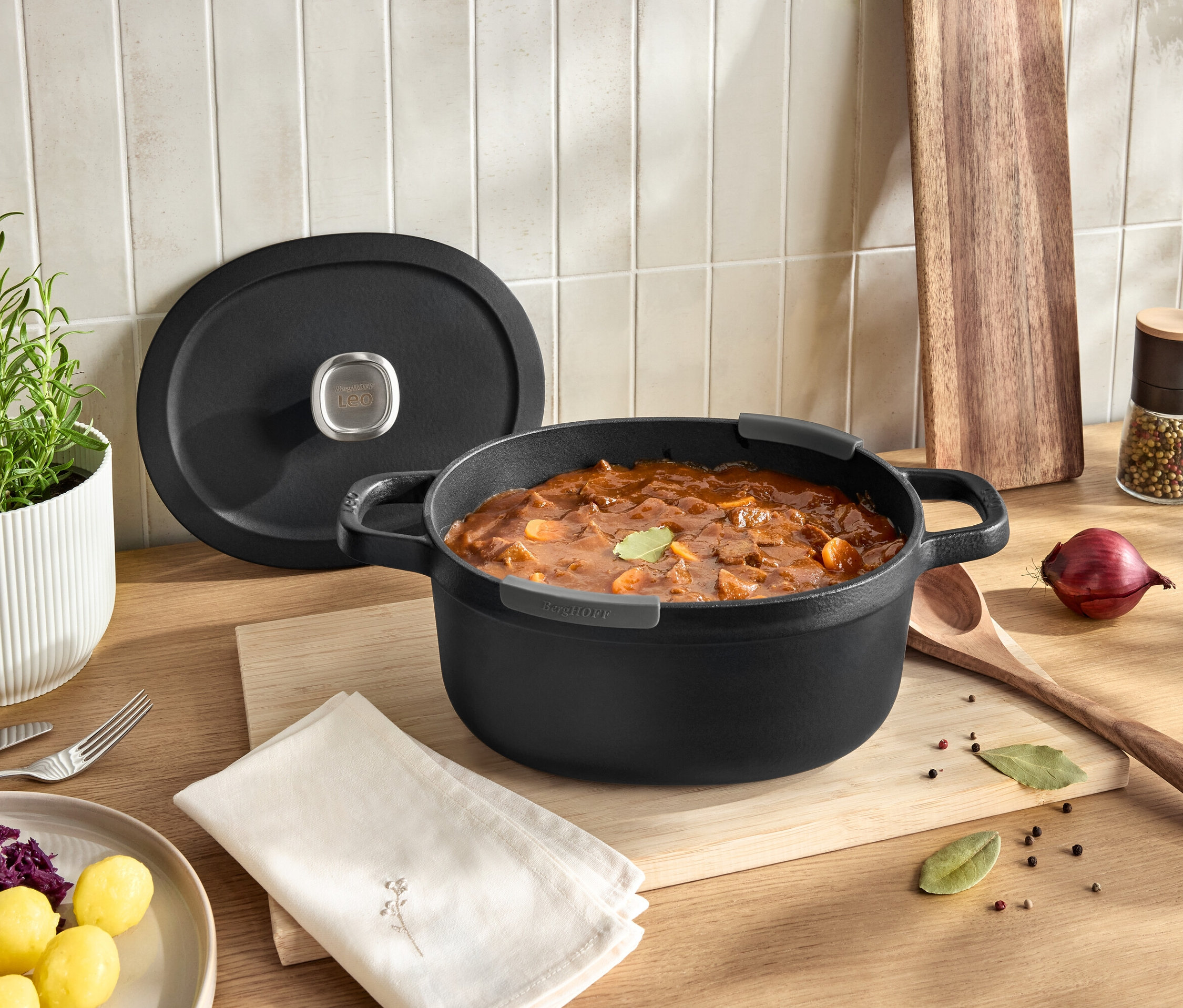 Schwarzer Topf mit Deckel voller Gulasch auf einem Holzbrett auf einem Tisch.