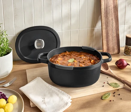 Schwarzer Topf mit Deckel voller Gulasch auf einem Holzbrett auf einem Tisch.