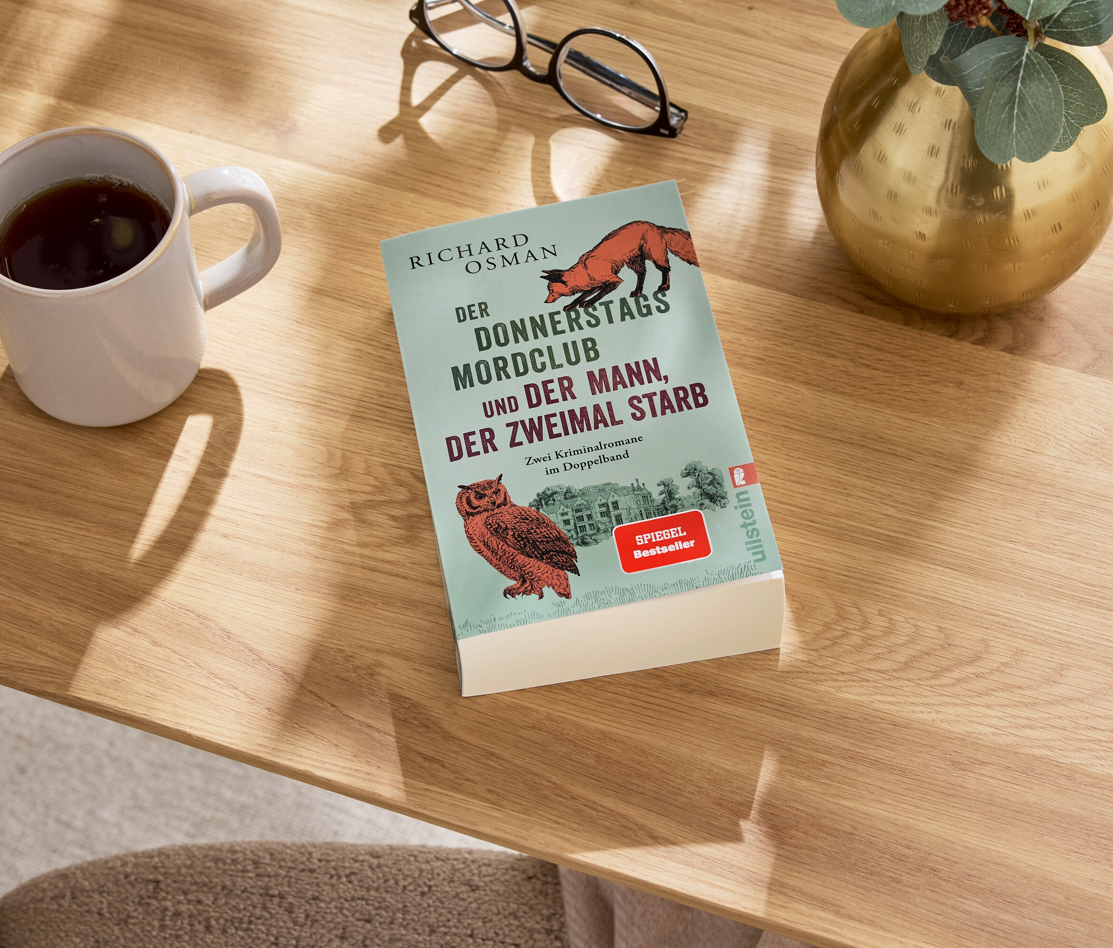 Auf einem Tisch liegen ein Buch, eine Tasse mit einem Getränk, eine Brille und eine Vase.