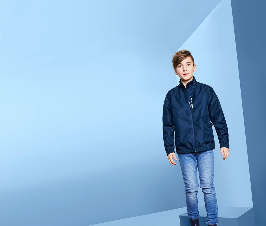 Junge steht auf blauen Würfeln und trägt eine Allwetterjacke im Bomberjacken-Stil.