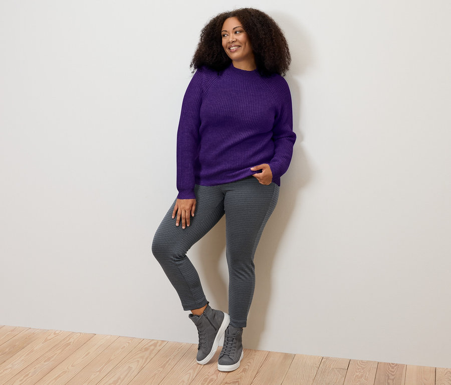 Frau posiert in lila Grobstrickpullover mit Stehkragen, grauen Jeggings und Turnschuhen.