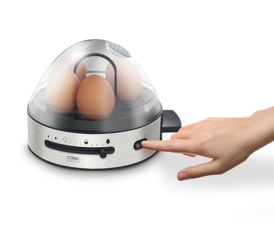 Eine Hand schaltet einen Eierkocher mit Eiern unter einer transparenten Abdeckung ein.