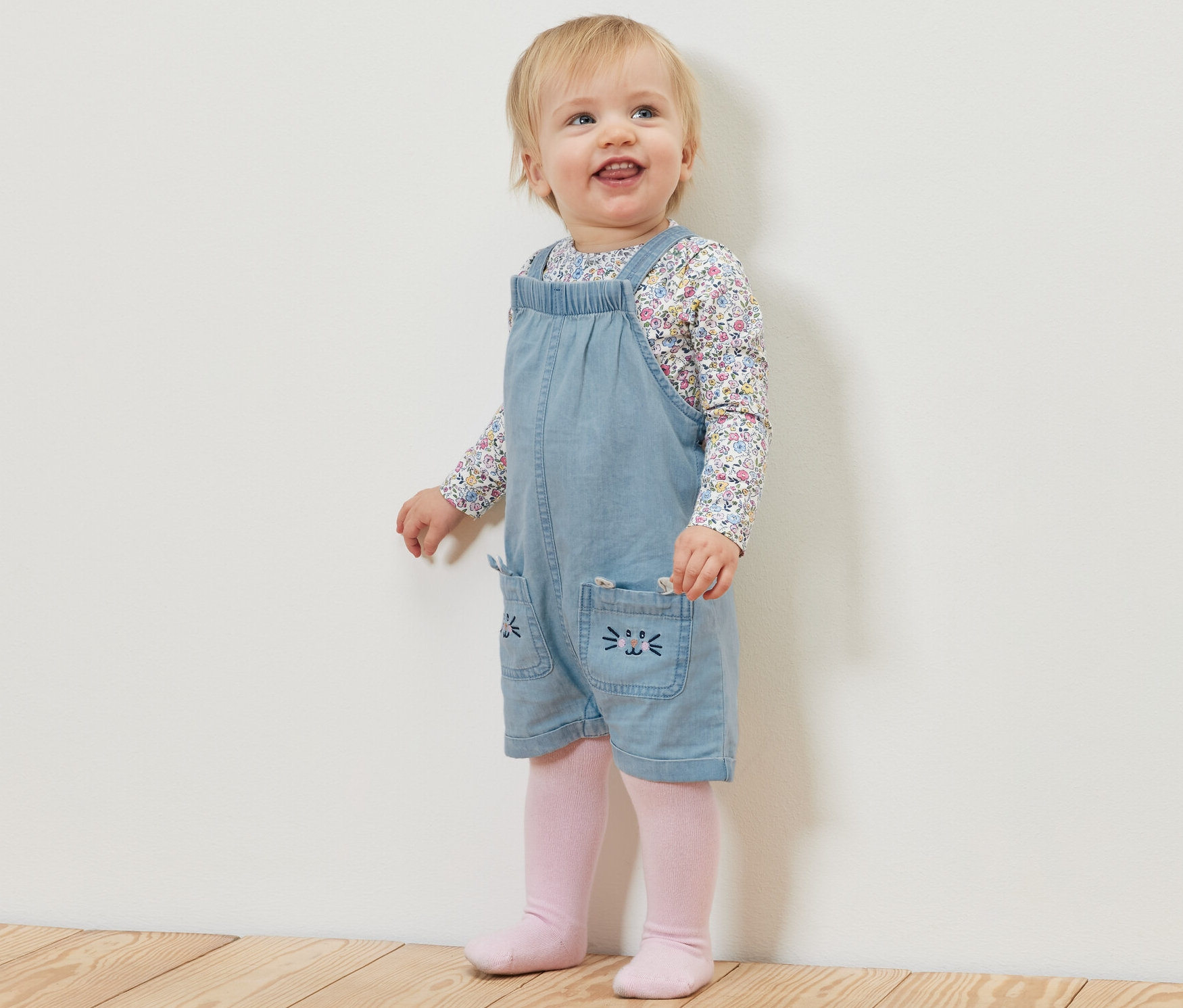 Kleinkind steht auf einem Holzboden und trägt eine Kinder Chambray-Latzhose und ein geblümtes Shirt.