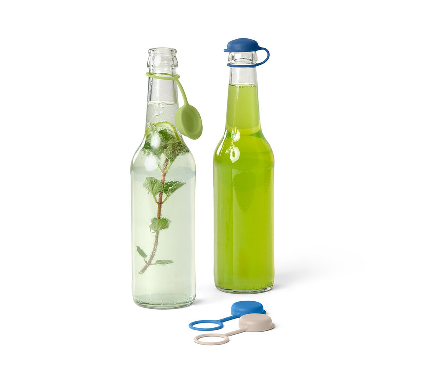 Zwei Glasflaschen, eine mit Minze und grünem Verschluss, die andere mit grünem Getränk und blauem Verschluss. Zwei weitere Flaschenverschlüsse liegen davor.