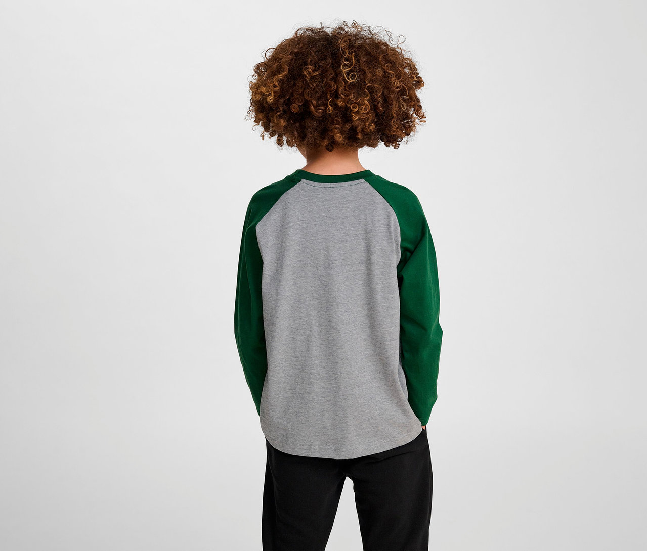 Junge mit lockigem Haar steht mit dem Rücken zur Kamera und trägt ein grünes HUMMEL HMLJR Raglan Loose T-Shirt.