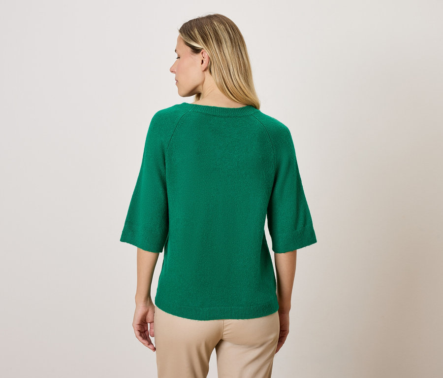 Frau trägt einen grünen Pullover mit Dreiviertelärmeln und eine beige Hose, mit dem Rücken zur Kamera.