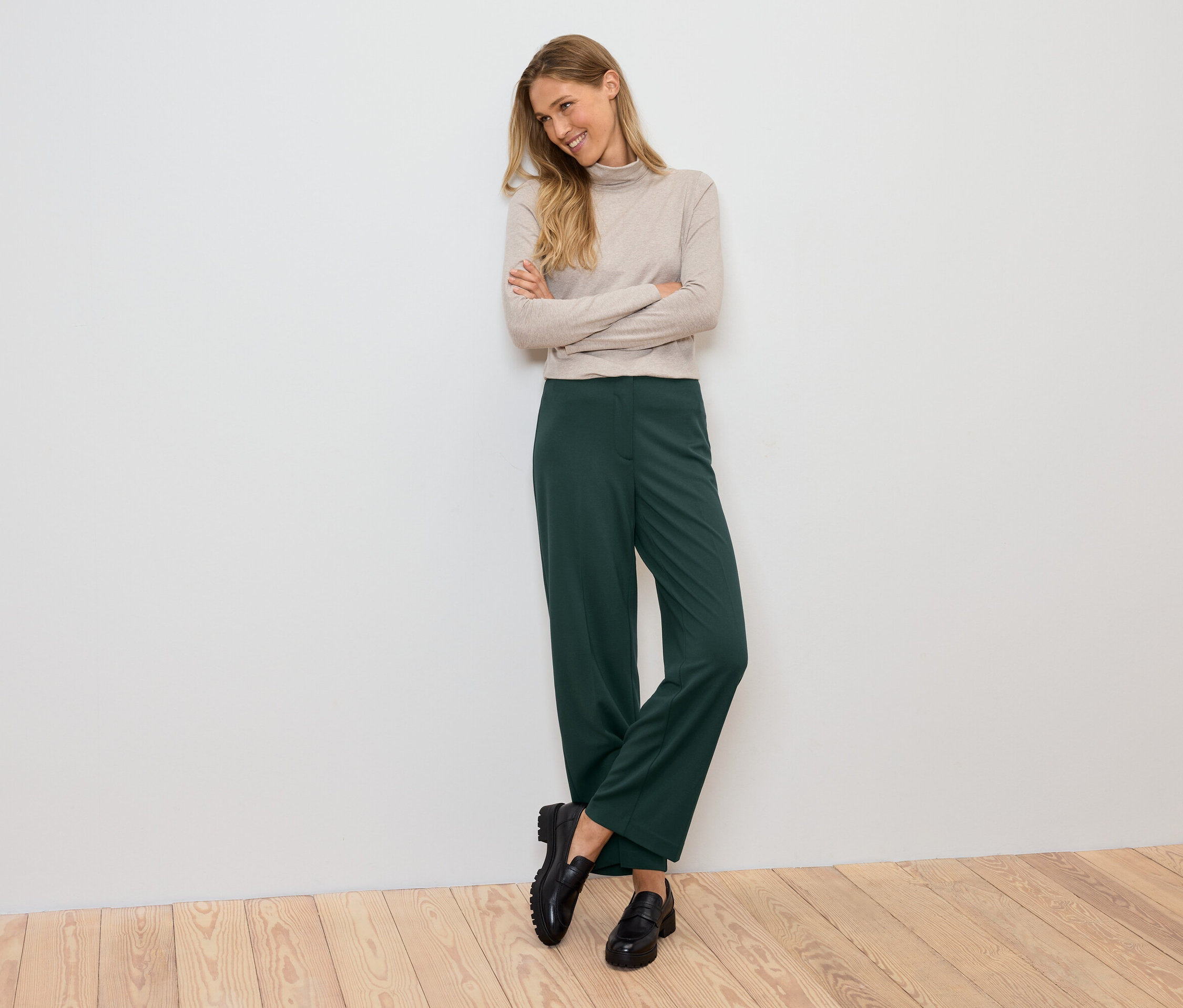 Eine lächelnde Frau posiert mit verschränkten Armen vor der Brust und trägt einen beigen Rollkragenpullover, eine grüne Hose und schwarze Schuhe.