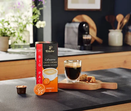 Eine Packung Cafissimo Caffe Crema Kapseln, ein Glas Kaffee und Kekse auf einem Tisch. Im Hintergrund eine Cafissimo pure+ black Kaffeemaschine.