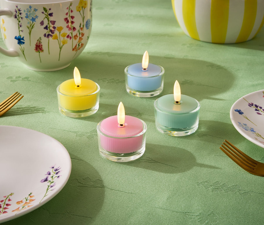 Auf einer grünen Tischdecke stehen vier kleine Kerzen in den Pastellfarben Gelb, Blau, Grün und Rosa. Dazu kommen ein Teller, eine Tasse und eine Schüssel mit Blumenmuster sowie zwei goldene Gabeln.