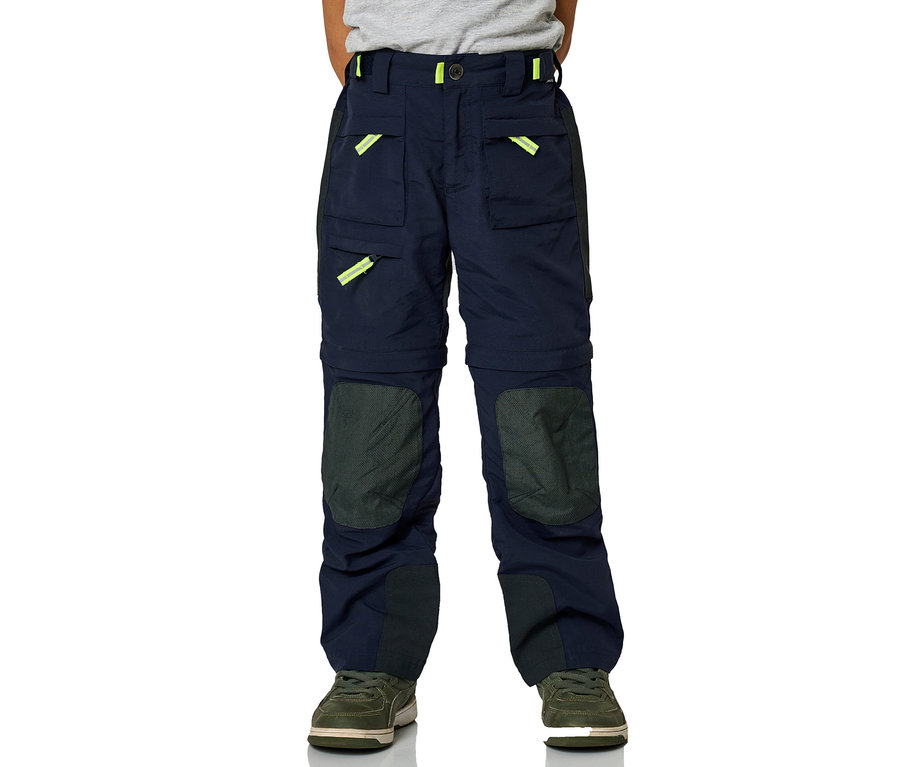 Ein Junge steht und trägt eine marineblaue Jako-O Zipp off Hose mit Taschen und grüne Schuhe.