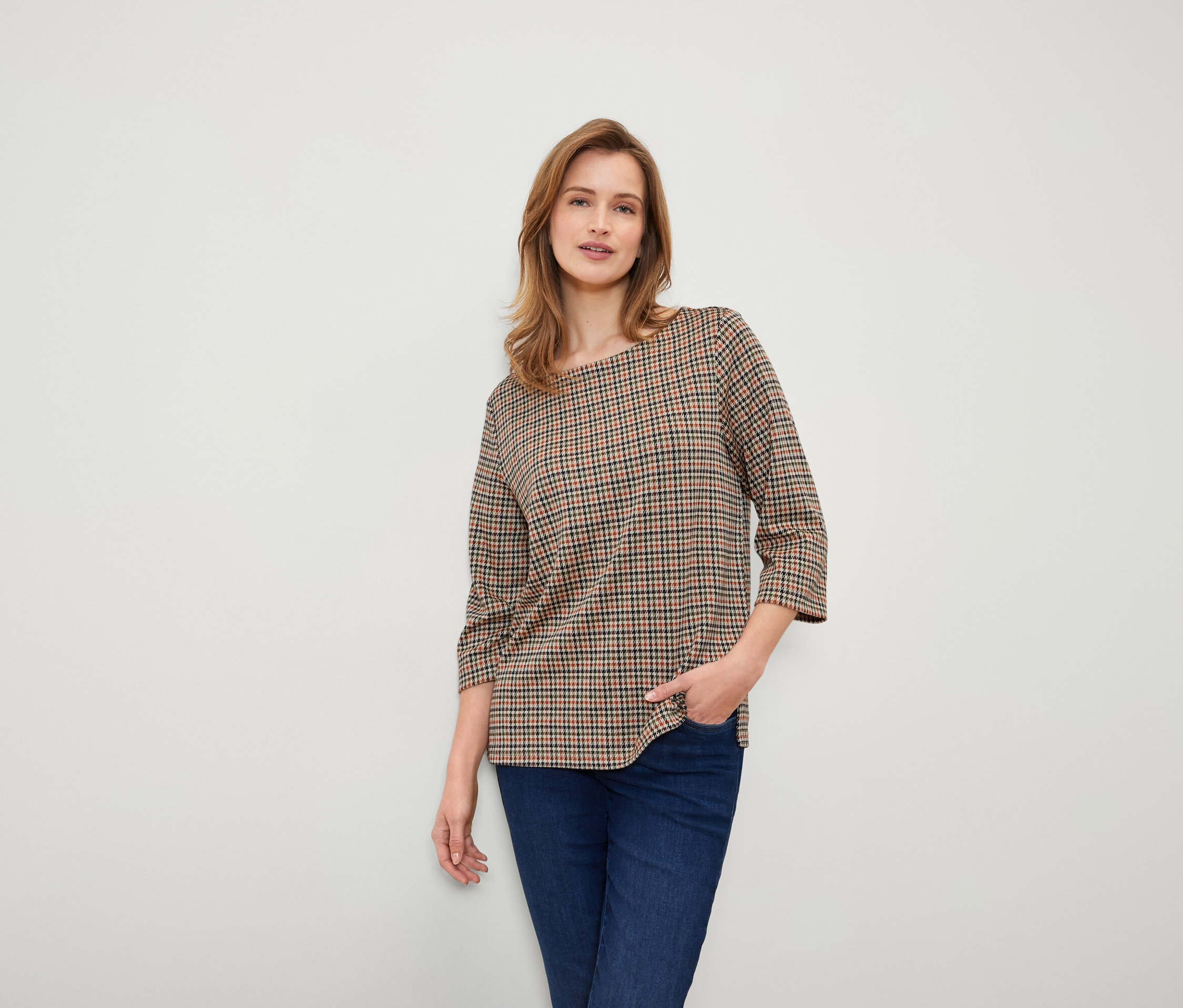 Frau mit braunen Haaren posiert in einer karierten braunen Bluse und dunkelblauen Jeans.