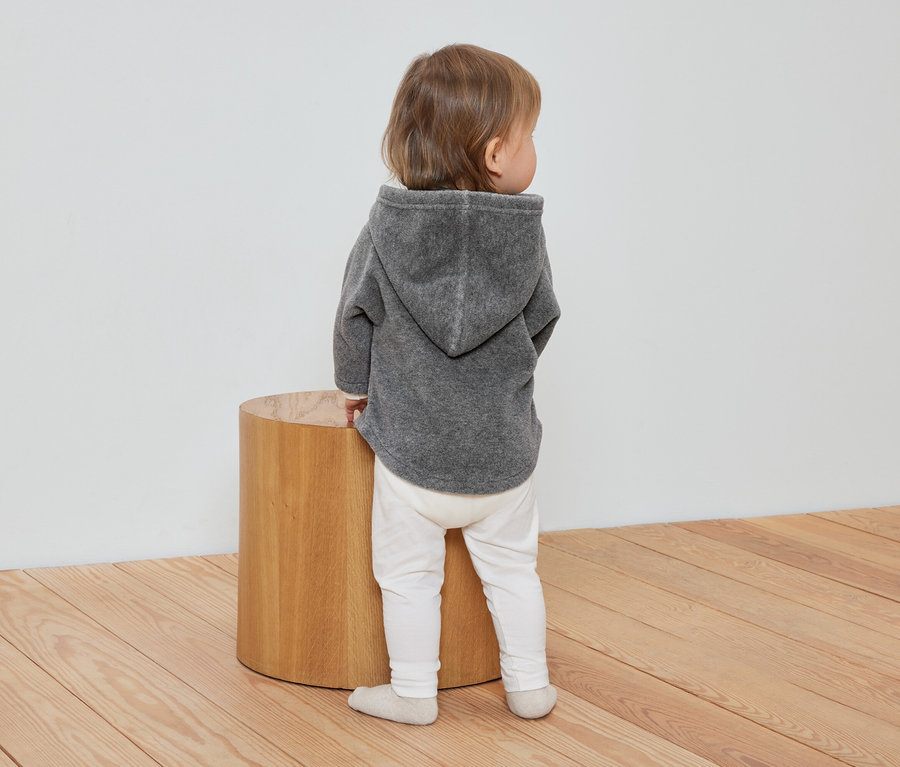 Kleinkind steht mit dem Rücken zur Kamera und trägt eine graue Baby-Fleecejacke.