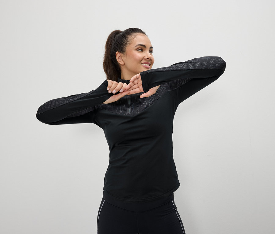 Eine lächelnde Frau in schwarzer Sportbekleidung dehnt sich vor einem weißen Hintergrund.