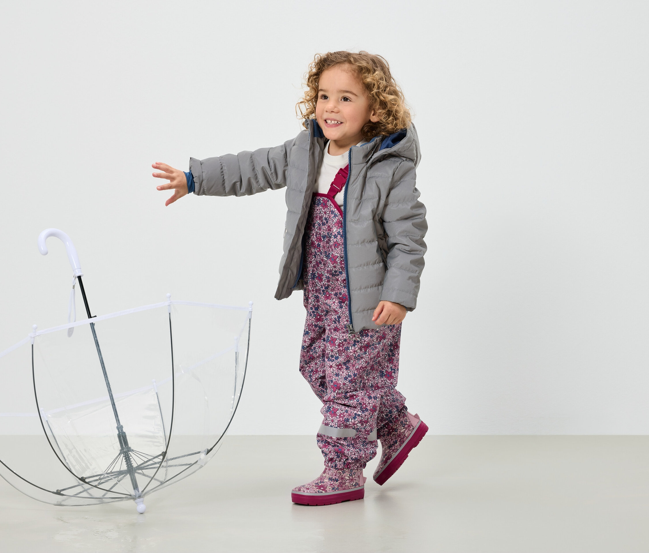 Ein lächelndes, lockiges Mädchen in grauer Jacke und geblümter Hose steht neben einem transparenten Regenschirm.