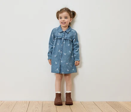 Ein kleines Mädchen steht auf einem Holzboden und trägt ein Jeanskleid mit Pferdeaufdruck und braune Stiefel.