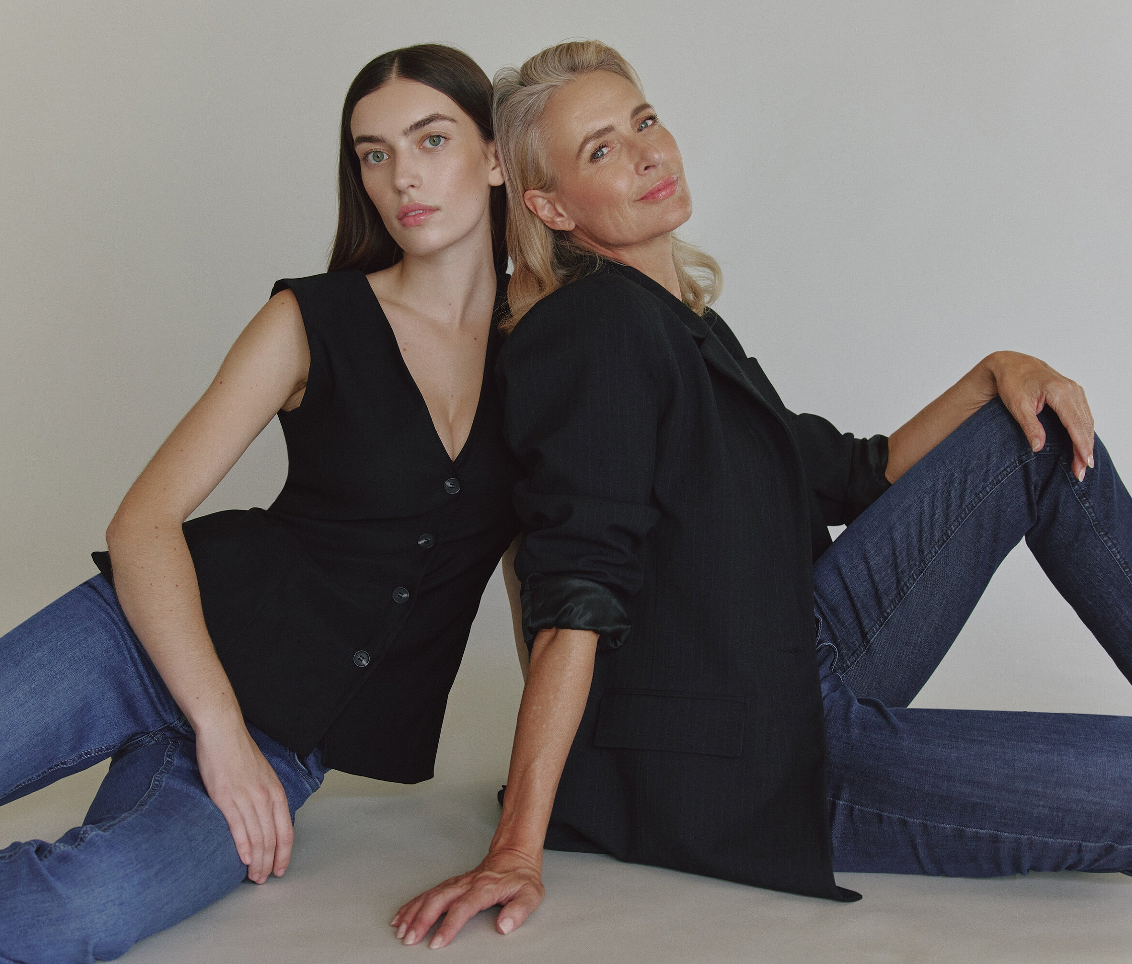 Zwei Frauen sitzen mit dem Rücken zueinander auf dem Boden. Eine trägt ein schwarzes Jackett und Jeans, die andere eine schwarze Weste und Jeans.