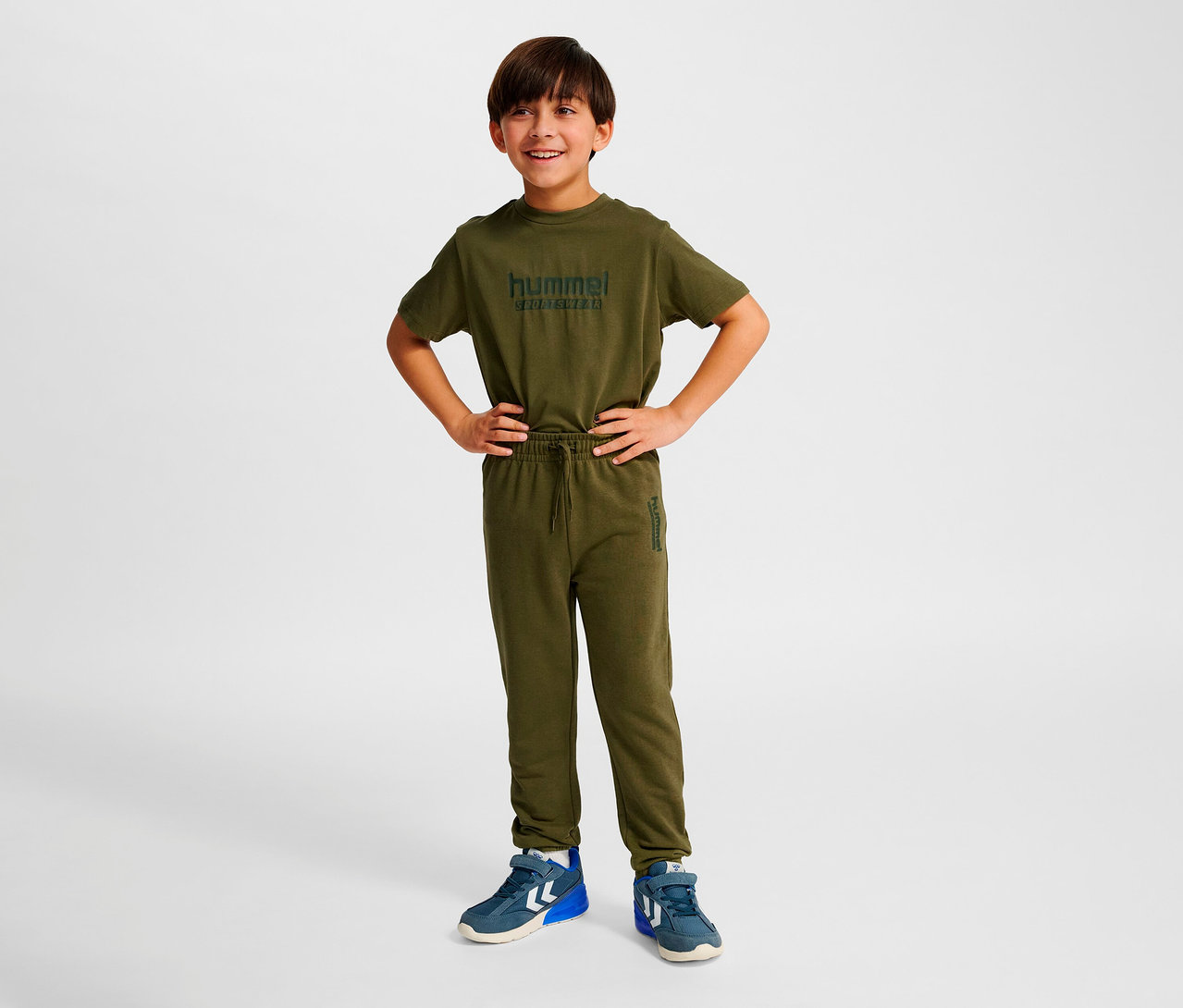 Junge steht mit Händen in den Hüften, trägt ein grünes Set mit Hummel HMLJR Base Regular Pants.