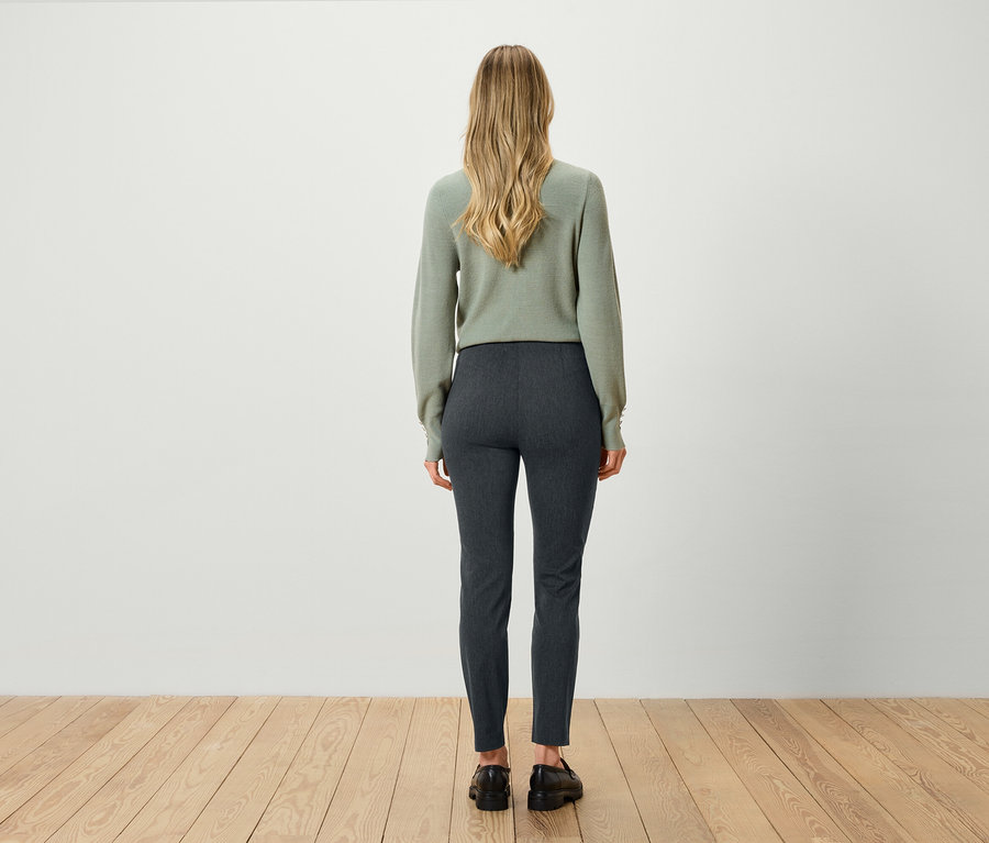 Rückansicht einer blonden Frau in einem grünen Pullover und grauen Hosen.