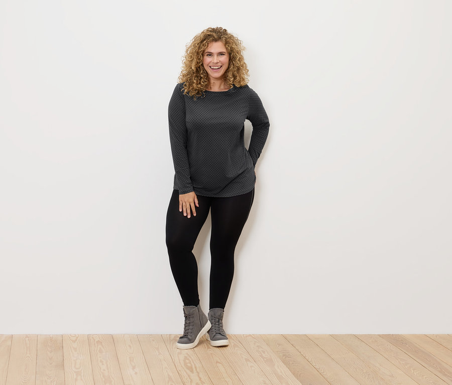 Frau mit lockigem Haar steht und lächelt, sie trägt schwarze Leggings, graue Schuhe und ein grau-schwarzes Shirt.