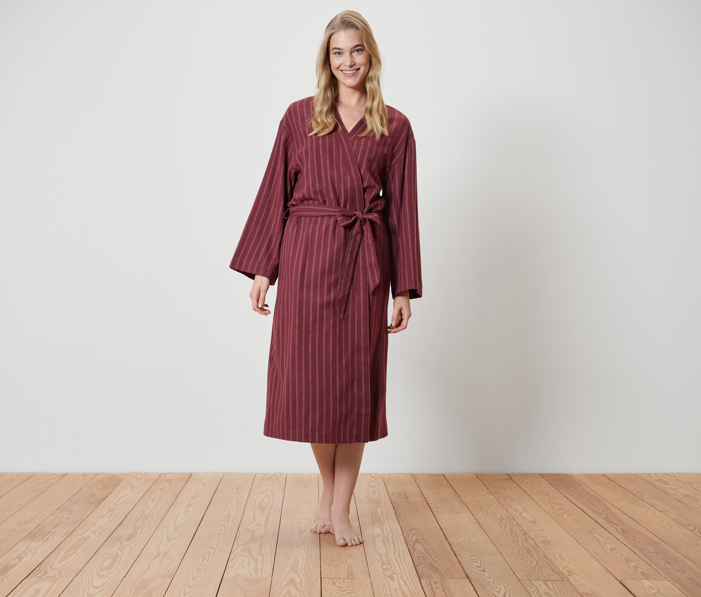 Eine blonde Frau steht auf einem Holzboden und trägt einen weinroten Bademantel mit Streifen.