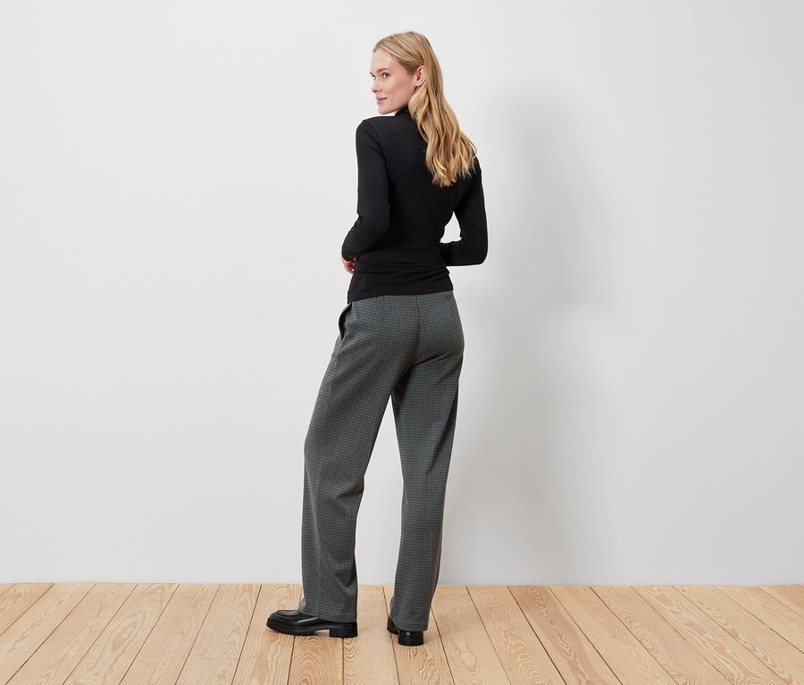 Blonde Frau in schwarzem Oberteil und grauer Hose blickt über die Schulter.