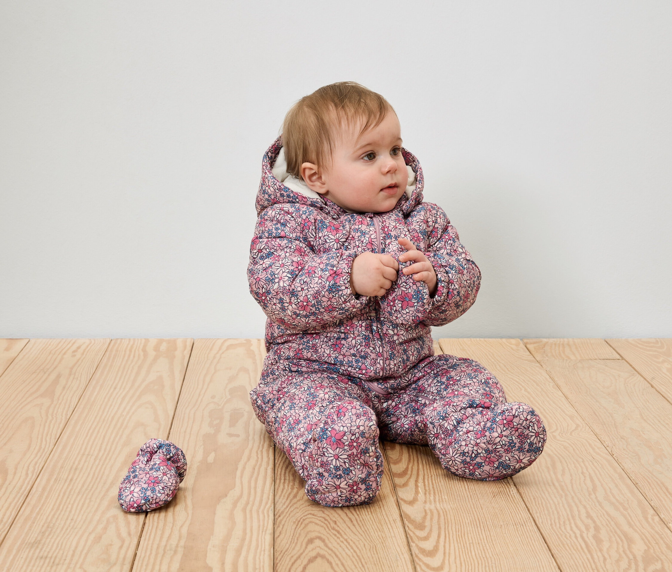Ein Baby-Mädchen sitzt auf einem Holzboden in einem geblümten Winteranzug mit Kapuze und einem Schuh.