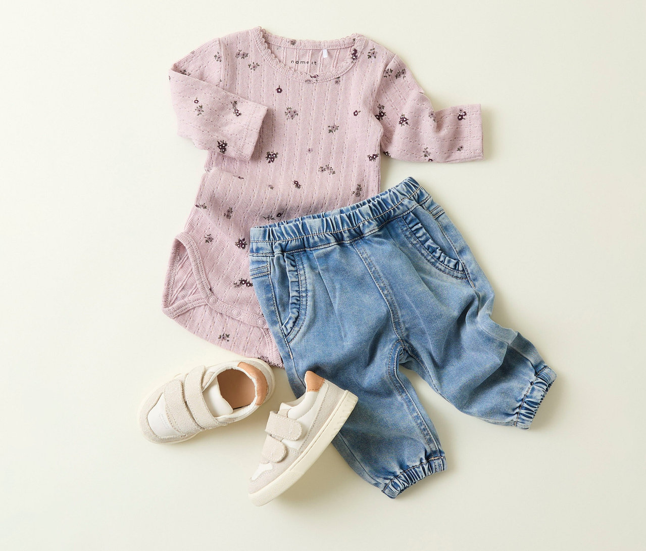 Ein rosafarbener Body mit Blumenmuster, eine NAME IT Baby-Jeans und beigefarbene Schuhe liegen auf einem hellen Hintergrund.
