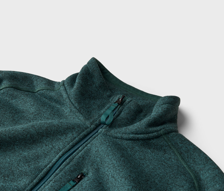 Nahaufnahme einer grünen Fleecejacke mit Reißverschluss und Stehkragen vor einem neutralen Hintergrund.