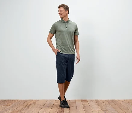 Ein Mann steht mit einer Hand in der Hosentasche vor einer weißen Wand, er trägt ein grünes Polohemd, blaue Shorts und schwarze Schuhe.