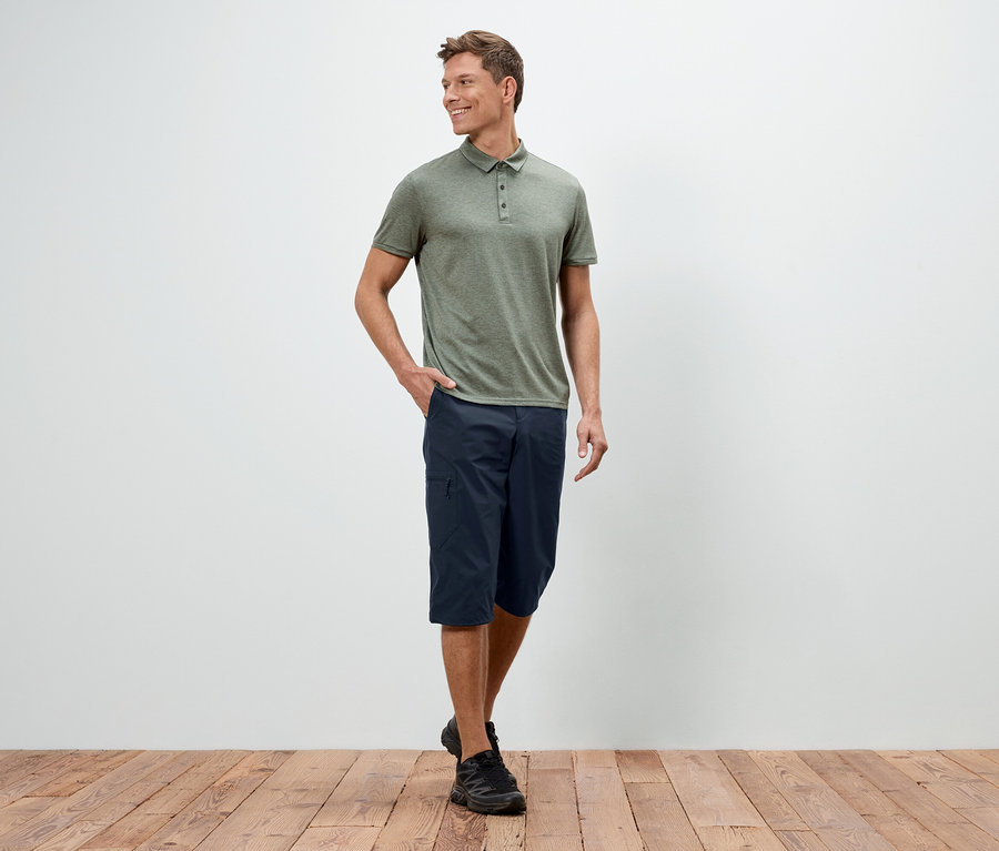 Ein Mann steht mit einer Hand in der Hosentasche vor einer weißen Wand, er trägt ein grünes Polohemd, blaue Shorts und schwarze Schuhe.