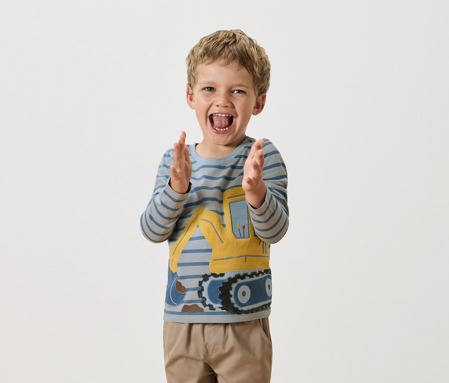 Ein Junge mit blonden Haaren klatscht in die Hände und trägt ein blau-gestreiftes Langarmshirt mit einem gelben Bagger darauf.