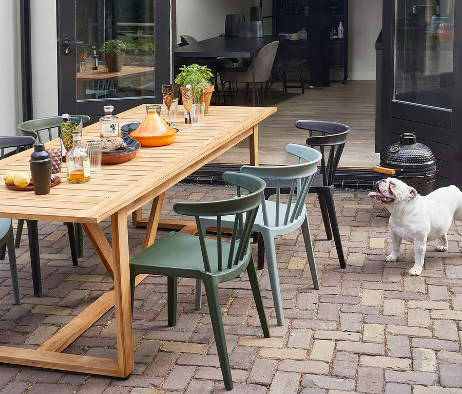 Ein Esstisch im Freien ist mit Getränken und Speisen gedeckt, umgeben von farbigen Stühlen und einem weißen Bulldog. Im Hintergrund steht ein Grill.