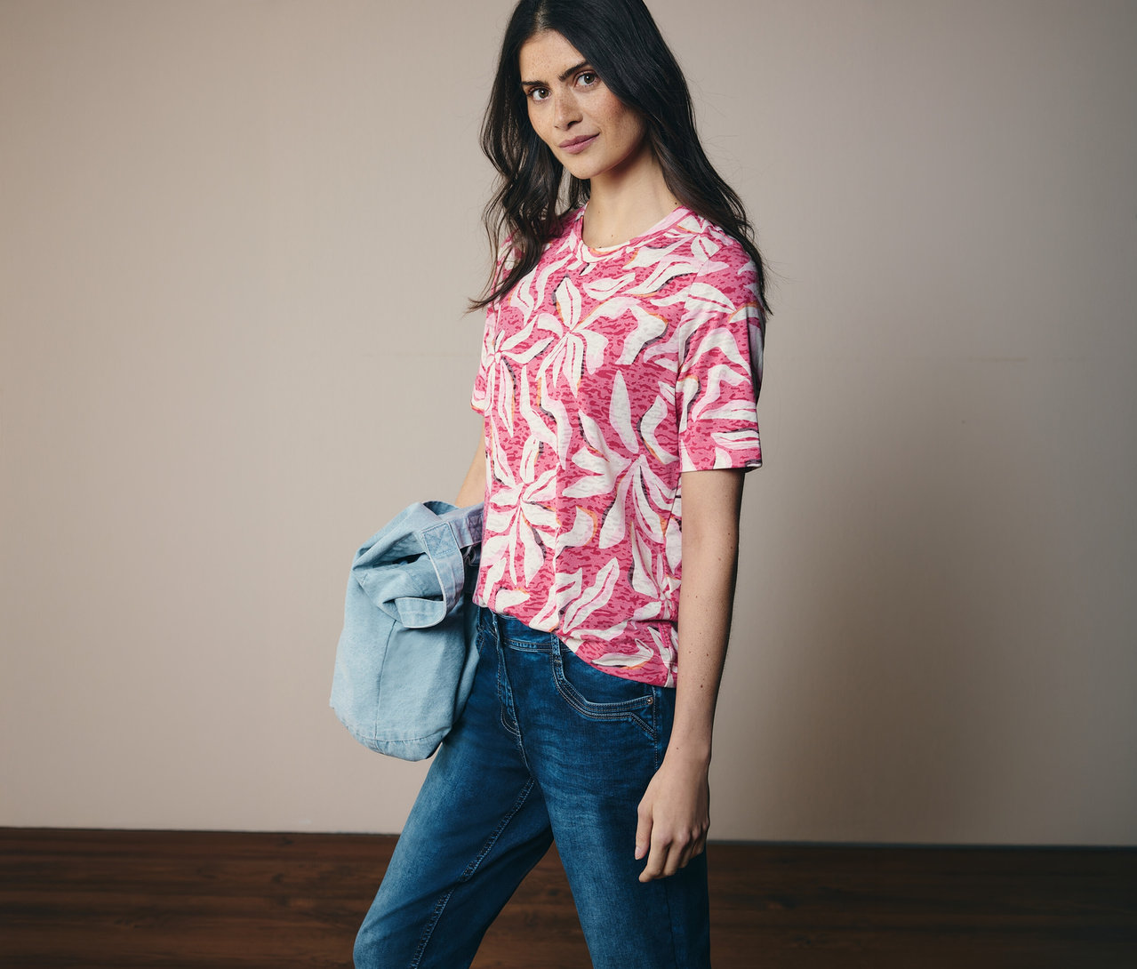 Frau trägt ein rosa CECIL Kurzarmshirt mit Muster und Jeans. Eine Jeansjacke hängt über ihrer Schulter.