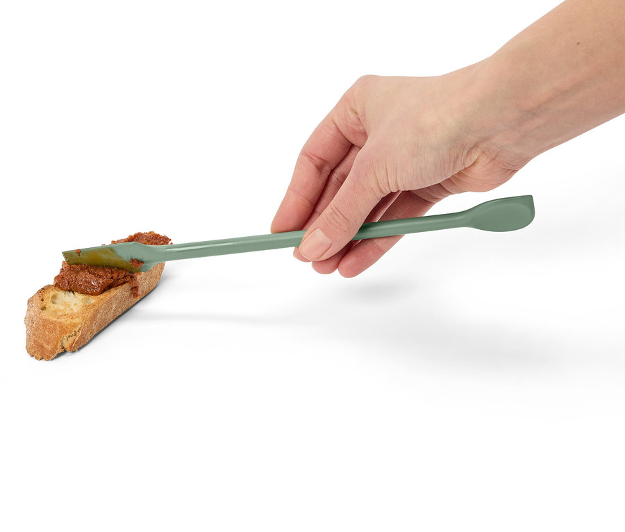 Eine Hand streicht mit einem grünen Spatel eine rote Paste auf eine Scheibe Brot.