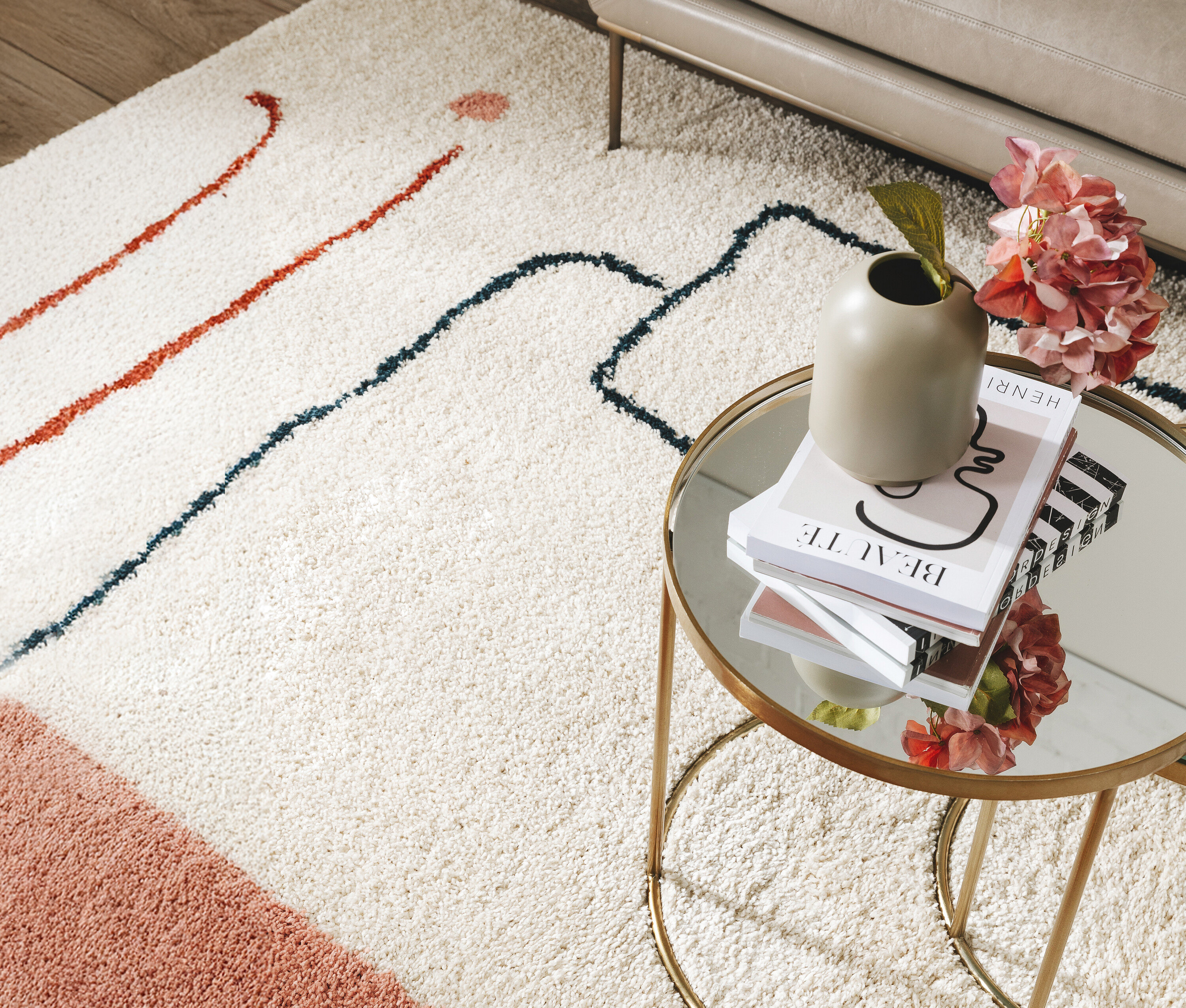 Ein beiger Teppich mit roten und blauen Linien liegt unter einem goldenen Beistelltisch mit Büchern, einer Vase und rosa Blumen.