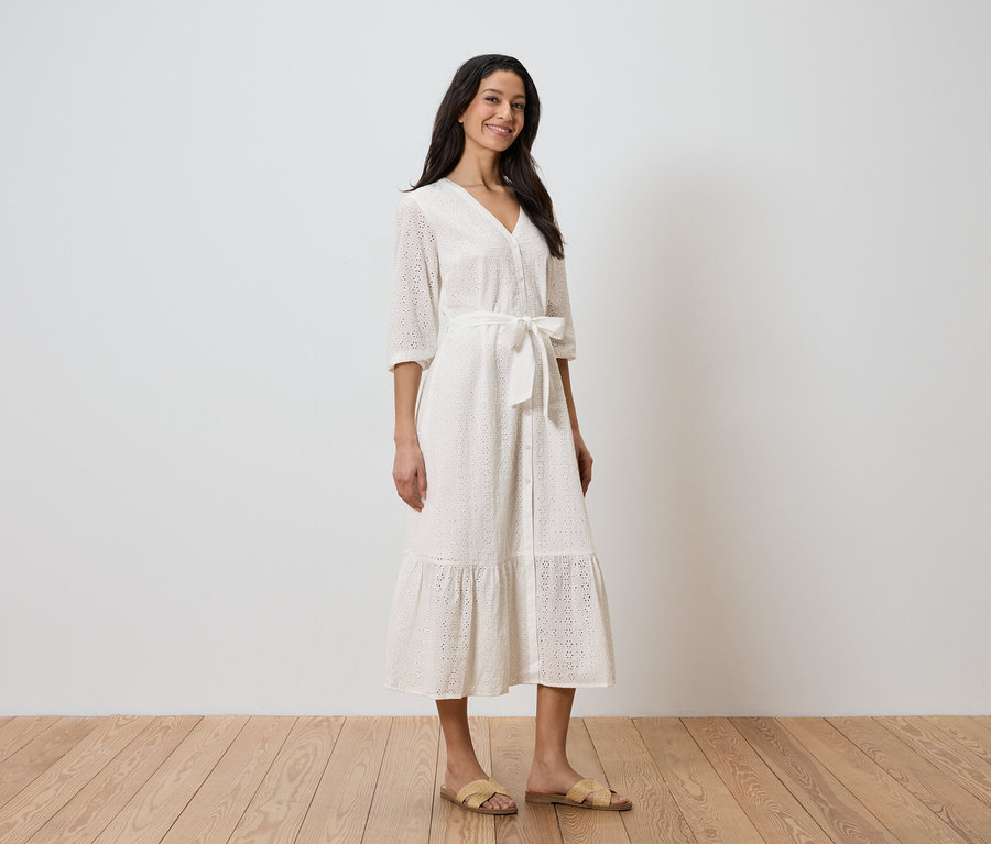 Eine Frau steht vor einer weißen Wand und trägt ein weißes Kleid mit Gürtel und Sandalen.