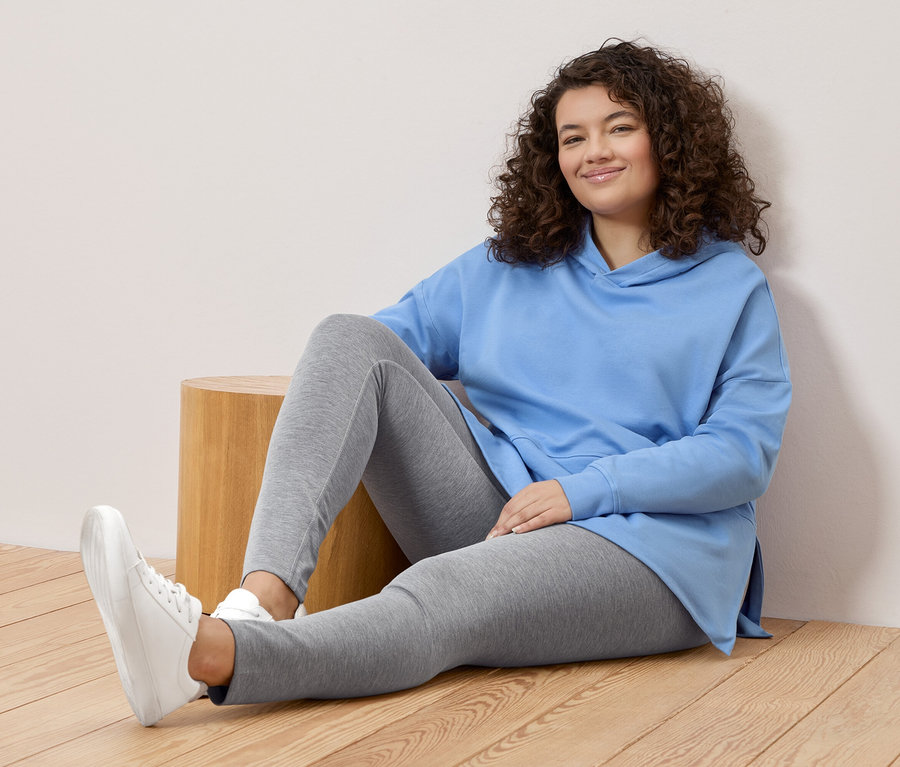 Frau mit lockigem Haar sitzt auf dem Boden, trägt einen hellblauen Hoodie, graue Leggings und weiße Turnschuhe.