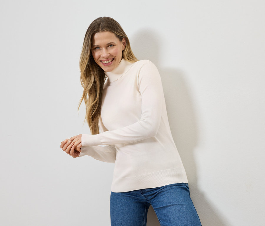 Frau mit blonden Haaren lächelt in einem cremefarbenen Rollkragenpullover und blauen Jeans vor einer weißen Wand.