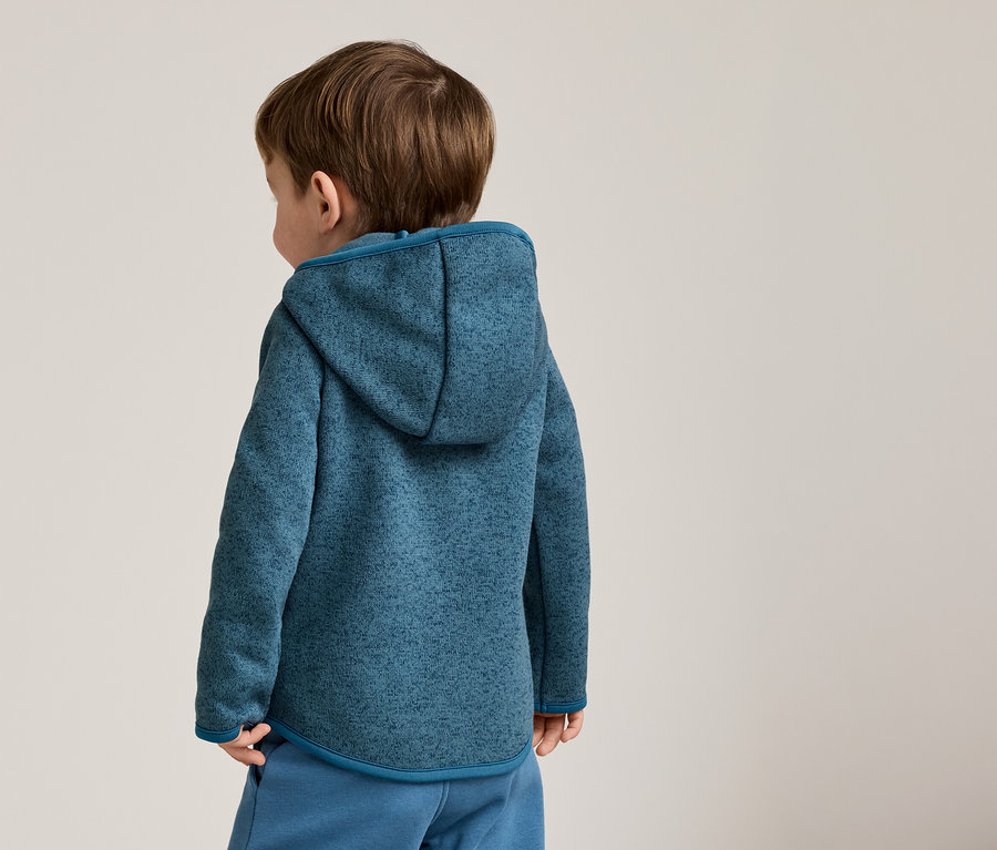 Rückansicht eines Kindes mit braunen Haaren, das einen blau melierten Kapuzenpullover und blaue Hosen trägt.