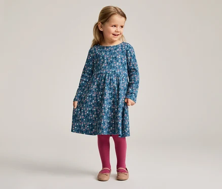 Ein kleines Mädchen mit blonden Haaren trägt ein blaues, langärmeliges Kleid mit Blumenmuster, dazu rosafarbene Strumpfhosen und Schuhe.