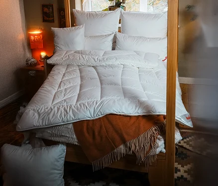 Bett mit weißer »ElbTraum« Winter-Faserdecke von Paradies, Normalgröße, mehreren Kissen und einer orangefarbenen Decke.