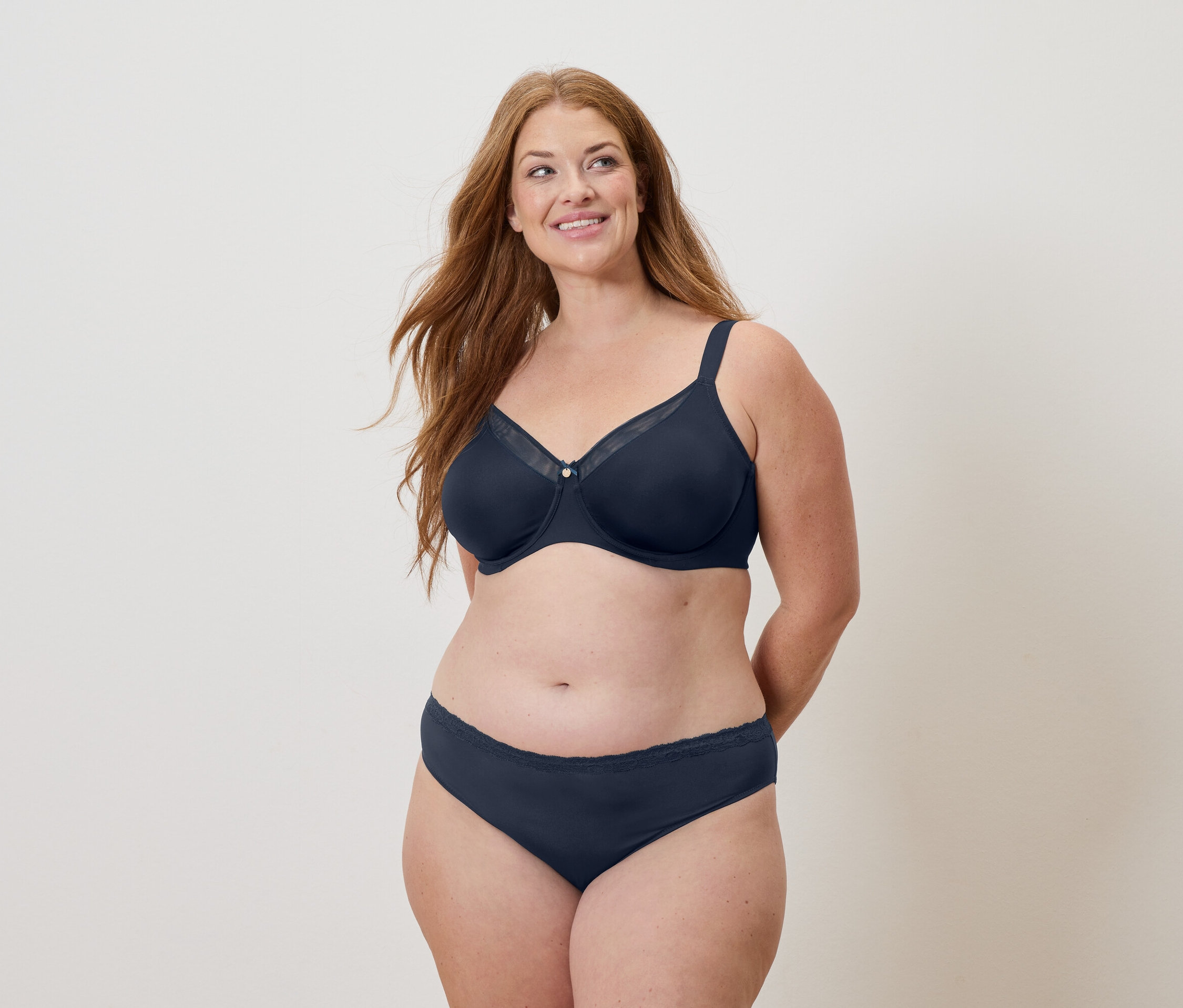 Frau mit langen roten Haaren trägt einen marineblauen BH und Slip und lächelt.