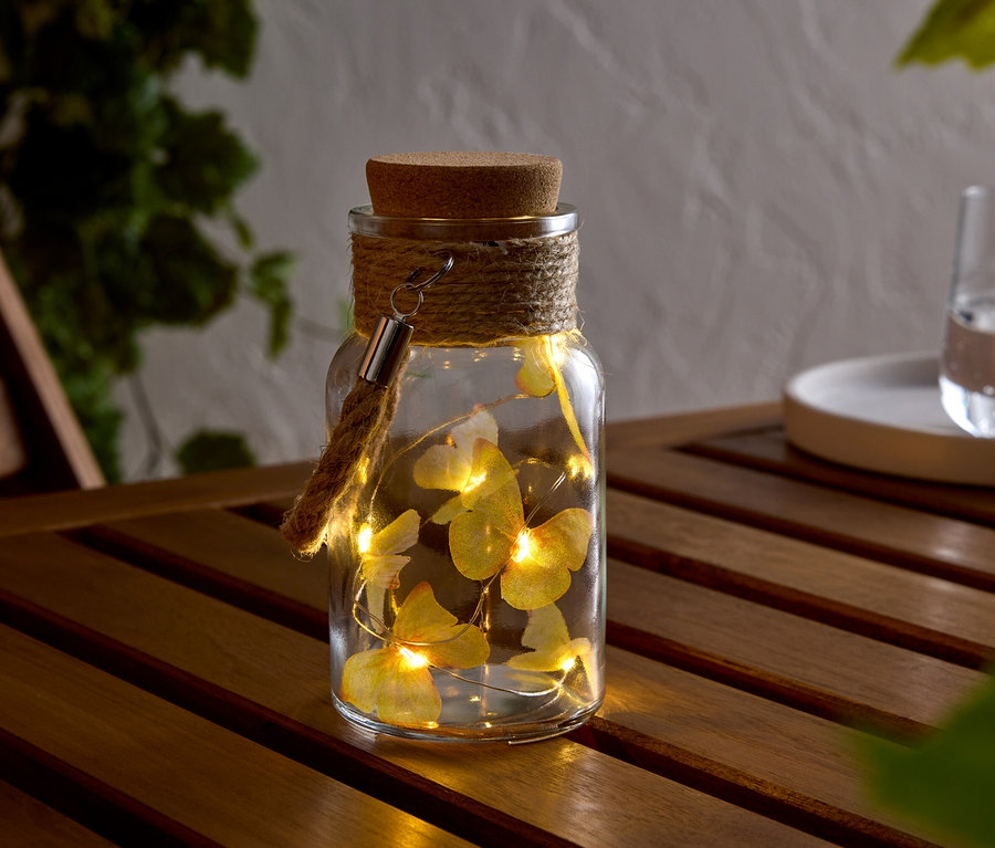 Einmachglas mit Korken, umwickelt mit einer Kordel, darin eine Lichterkette mit Schmetterlingen, auf einem Holztisch.