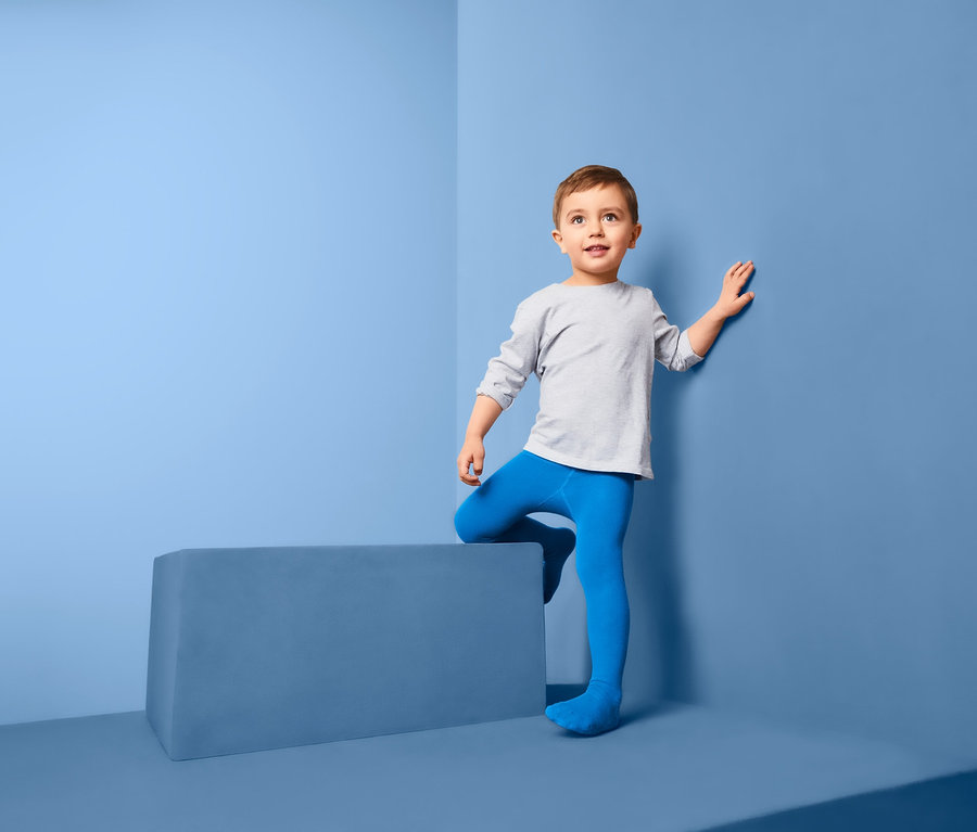 Ein Junge in grauem Hemd und blauer Strumpfhose, 3 Paar, steht in der Ecke eines blauen Zimmers.