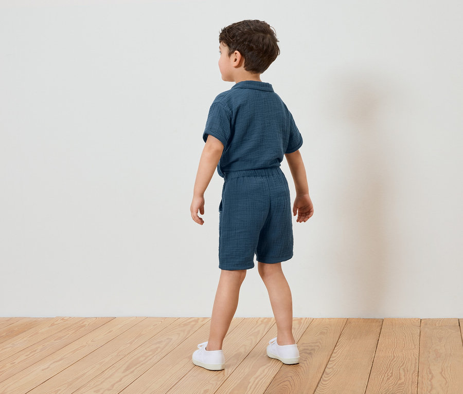 Junge von hinten in blauem Musselin-Shorts-Set und weißen Schuhen, vor einer weißen Wand und Holzboden.