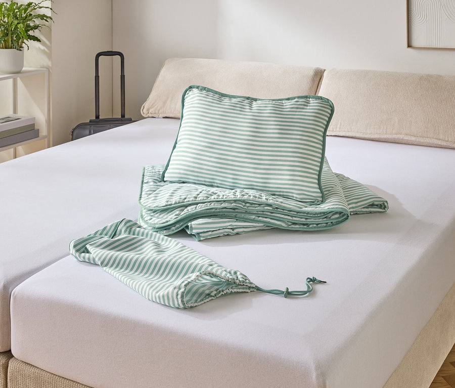 Ein Bett mit weißem Spannbettlaken, darauf ein gestreiftes Kissen, eine Decke und ein Beutel.